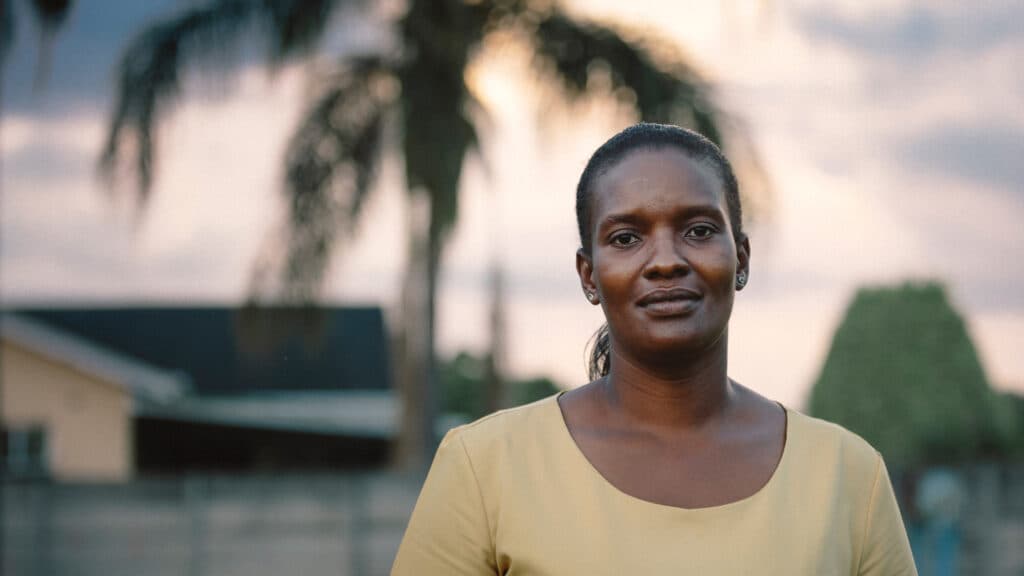 En kvinna tittar in i kameran. I bakgrunden ett hus och en palm.