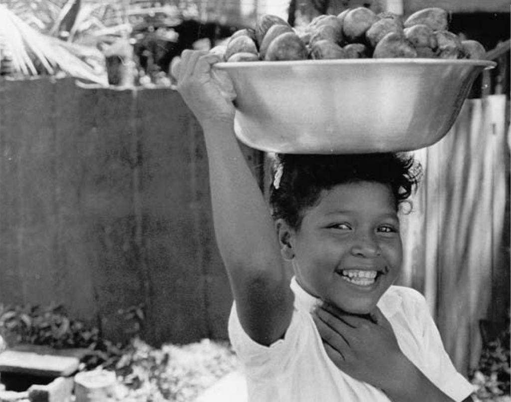 Ett svartvitt foto av ett leende barn som bär ett kärl på huvudet.