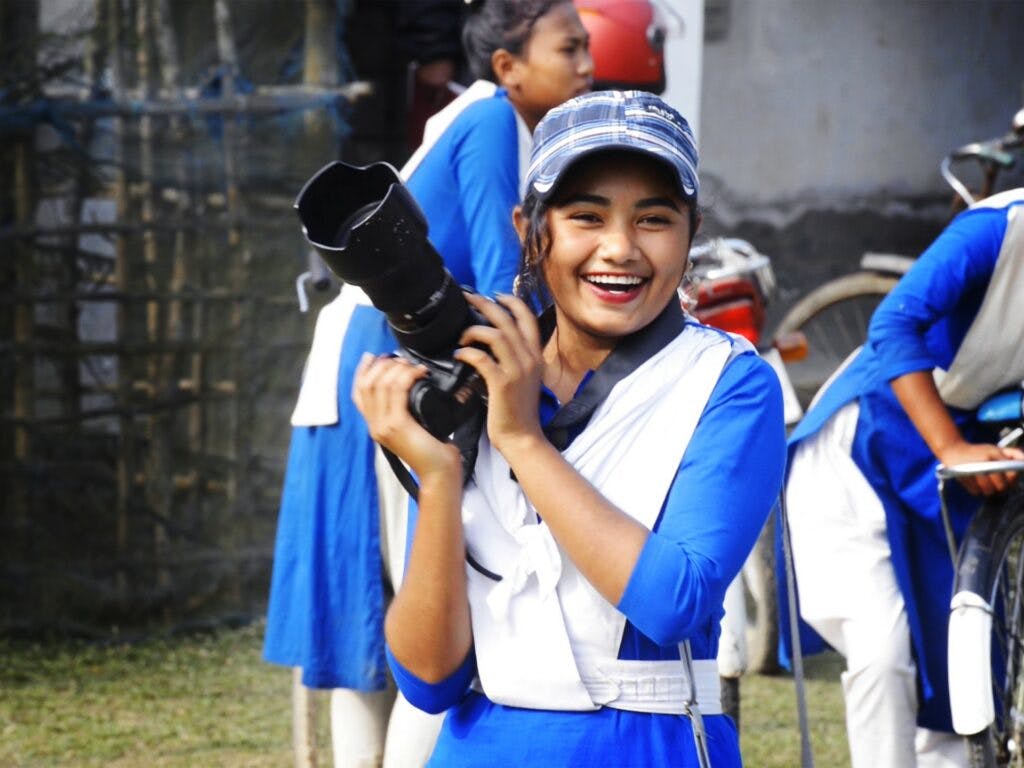 En flicka i skoluniform håller i en stor kamera och ler.