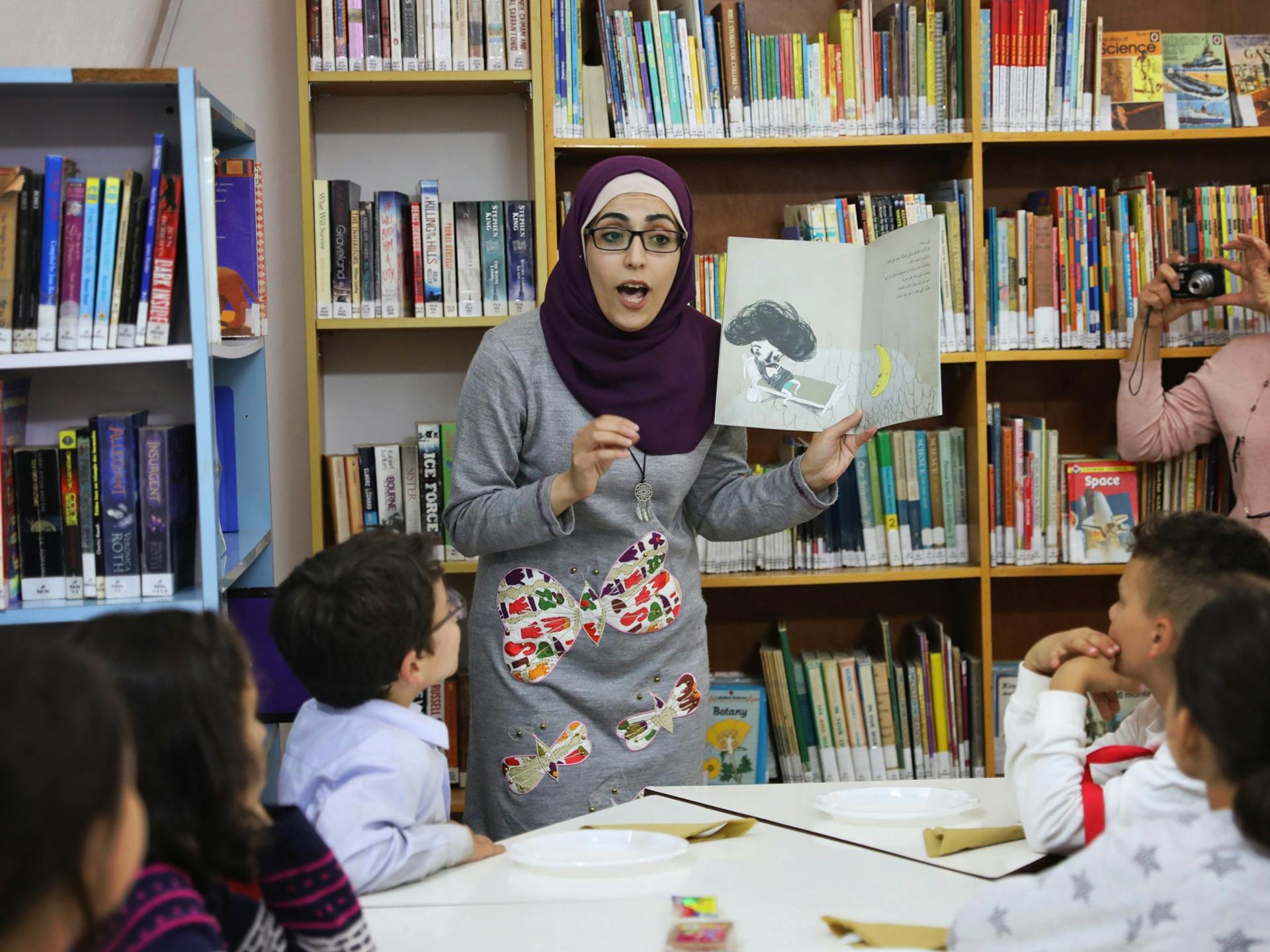 En kvinna med glasögon och hijab håller en bilderbok i handen och gestikulerar inför en grupp med barn.