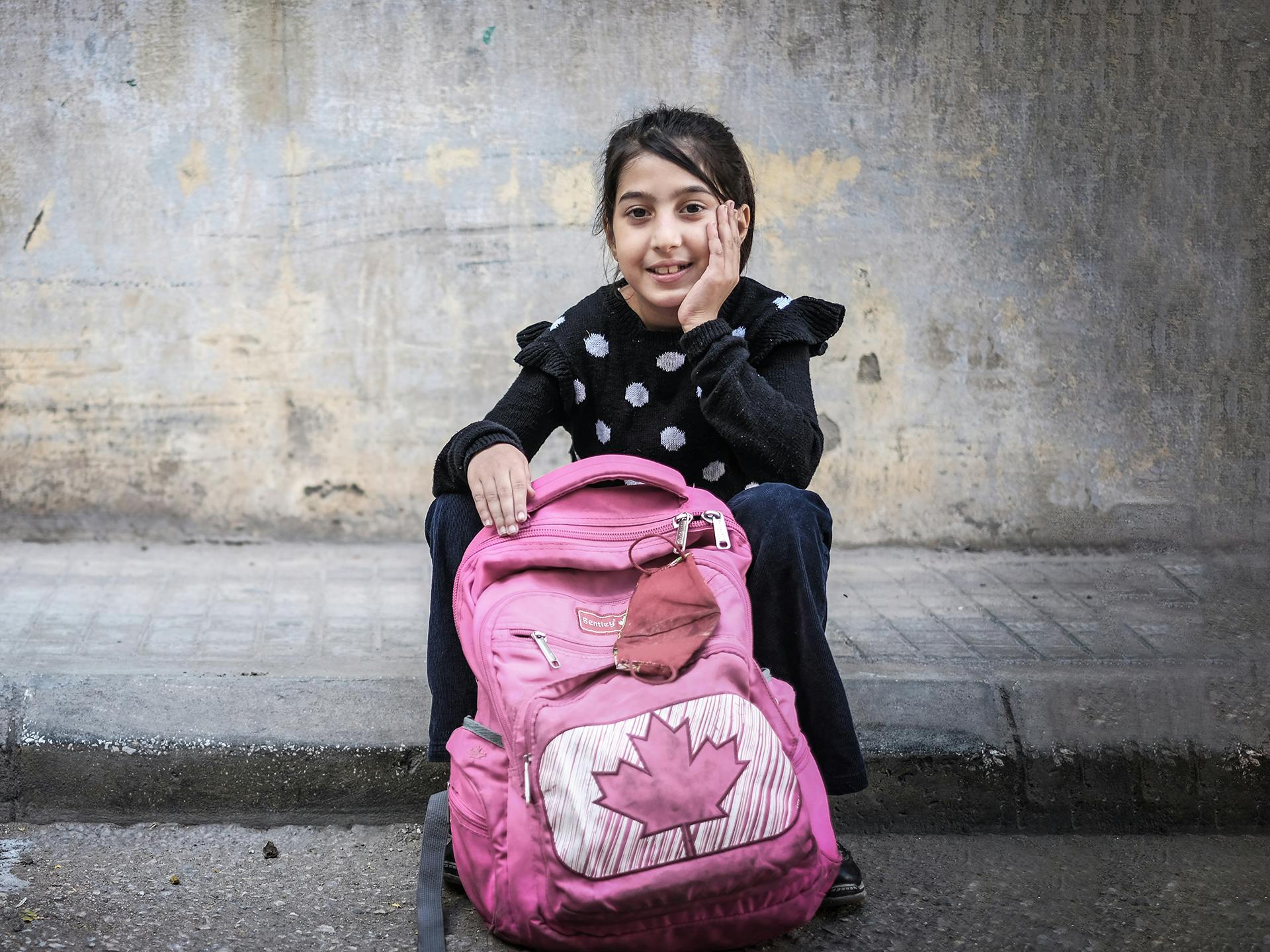 En flicka sitter på en trottoarkant med en rosa ryggsäck framför sig.