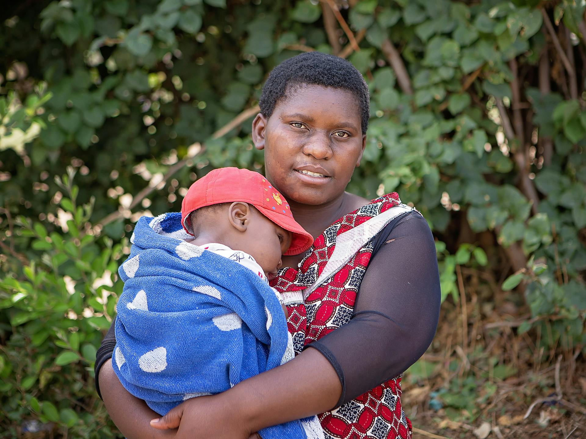 En kvinna håller en bebis. I bakgrunden syns ett stort buskage.