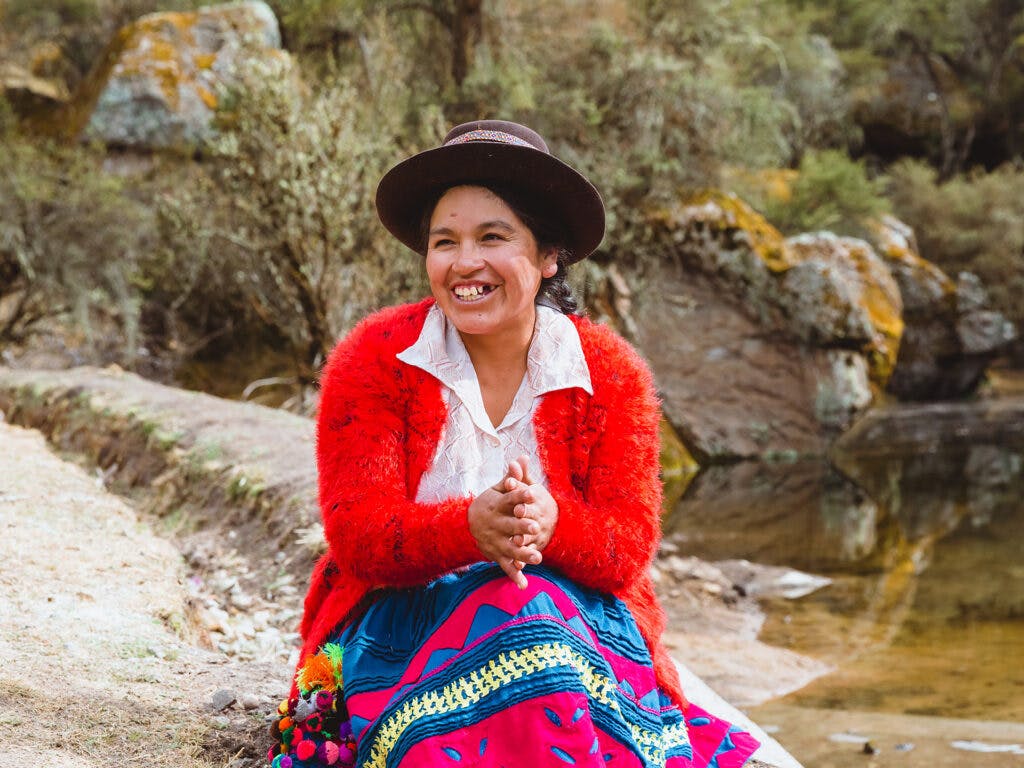 En kvinna i hatt och färgglad kjol sitter på en klippa och ler.