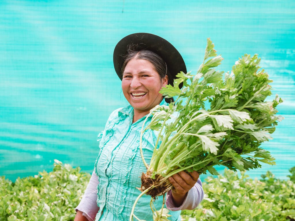 En kvinna med hatt och turkos blus håller upp en knippe grönsaker i handen.