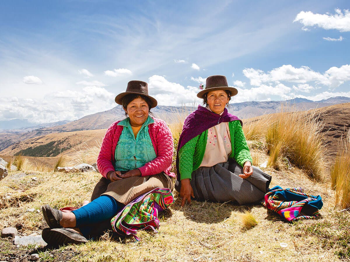 Två kvinnor med hattar och färgglada kläder sitter på marken i ett kargt landskap.