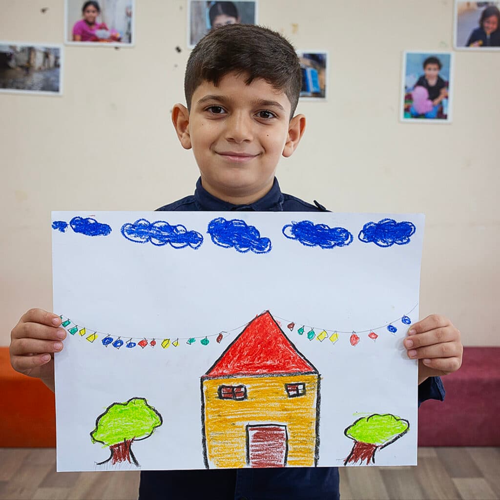 Foto: Samar Abu Elouf En pojke håller upp en teckning. På teckningen är ett gult hus med träd runt omkring.