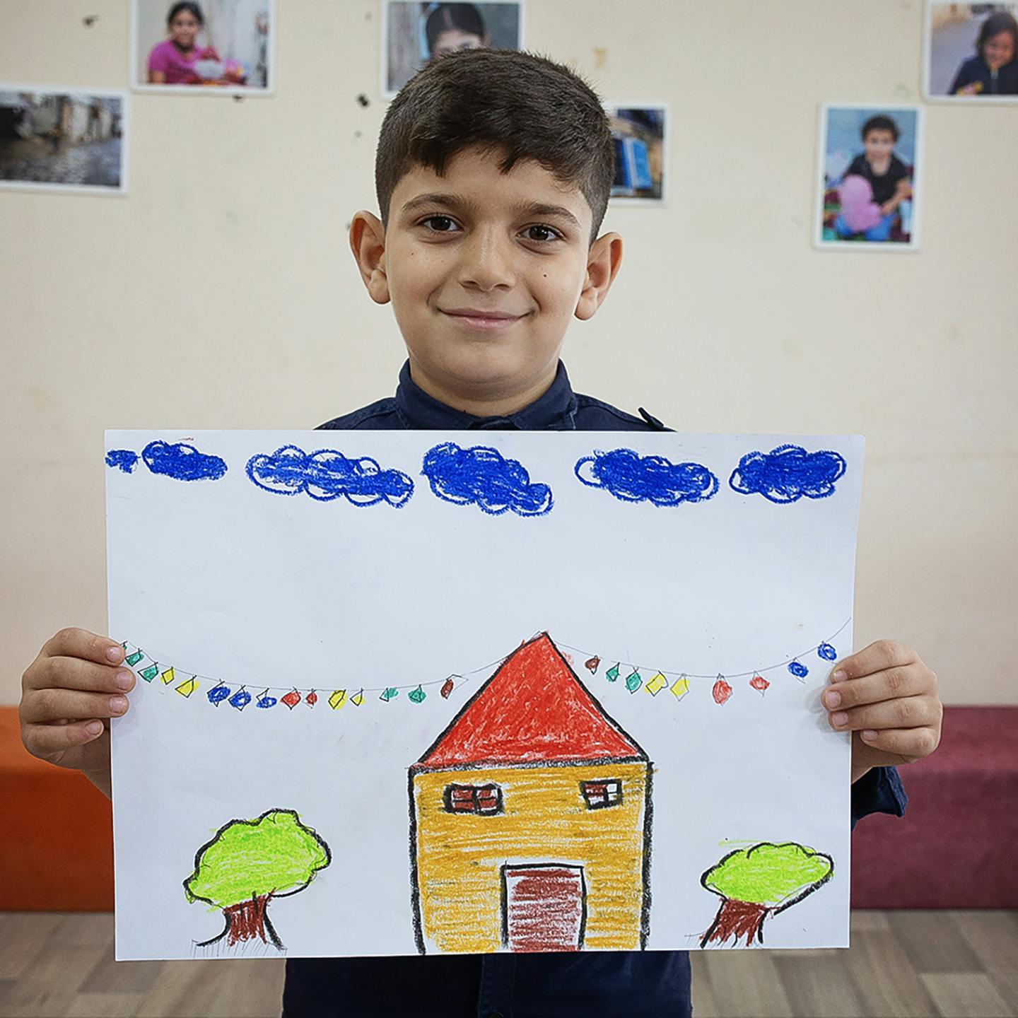En pojke håller upp en teckning. På teckningen är ett gult hus med träd runt omkring.