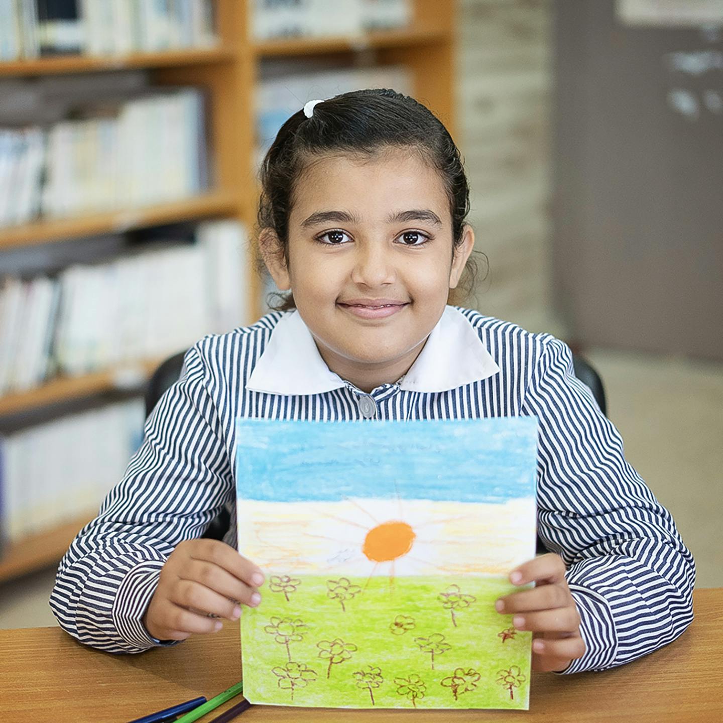 En flicka håller upp en teckning. Teckningen är en sol med himmel ovanför och gräs nedanför.