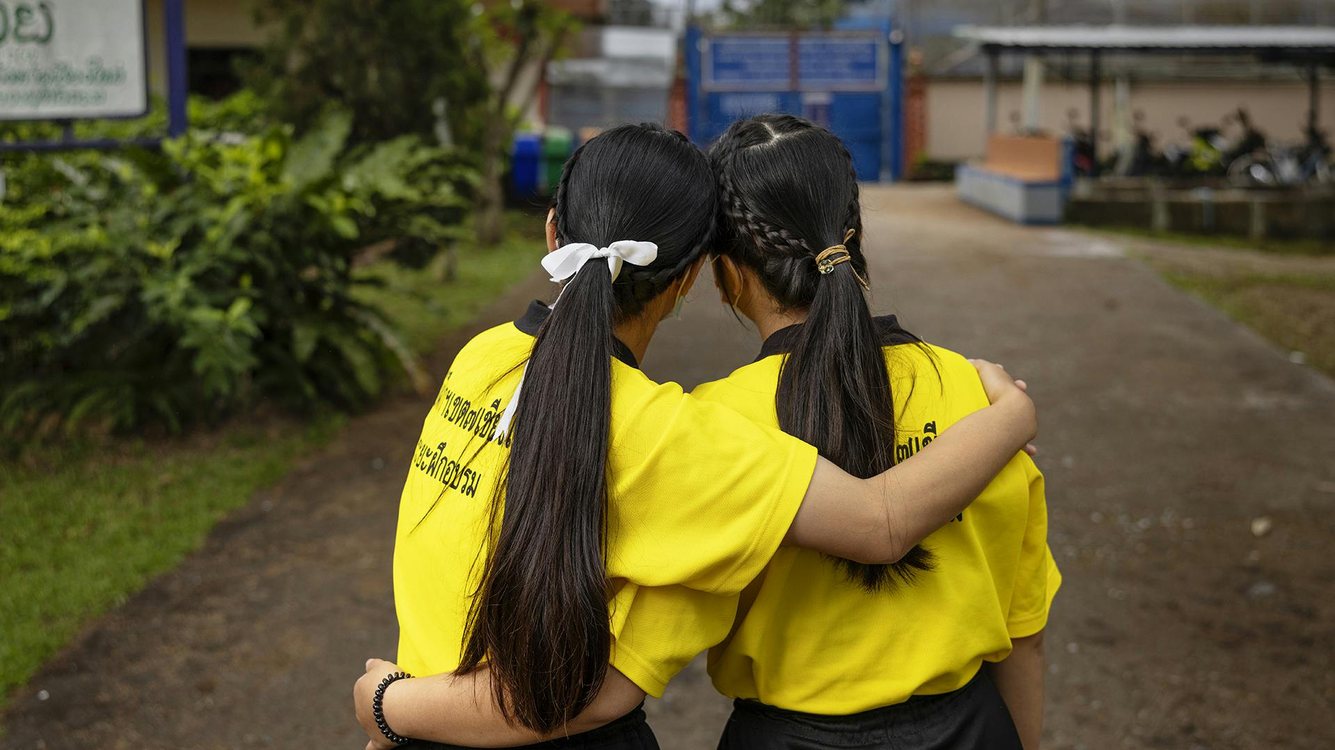 Två unga kvinnor står med ryggarna mot kameran och håller om varandra. De har likadana gula tröjor på sig och långt mörk hår i hästsvans.