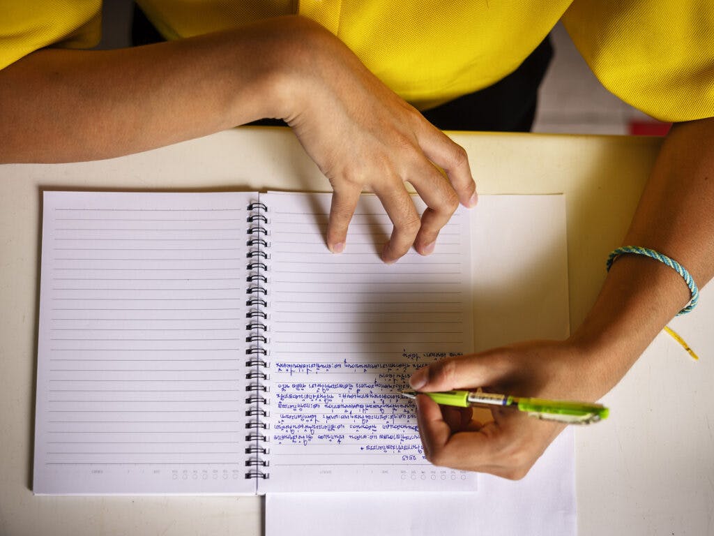Två händer som skriver i en skrivbok på thai, sett ovanifrån.