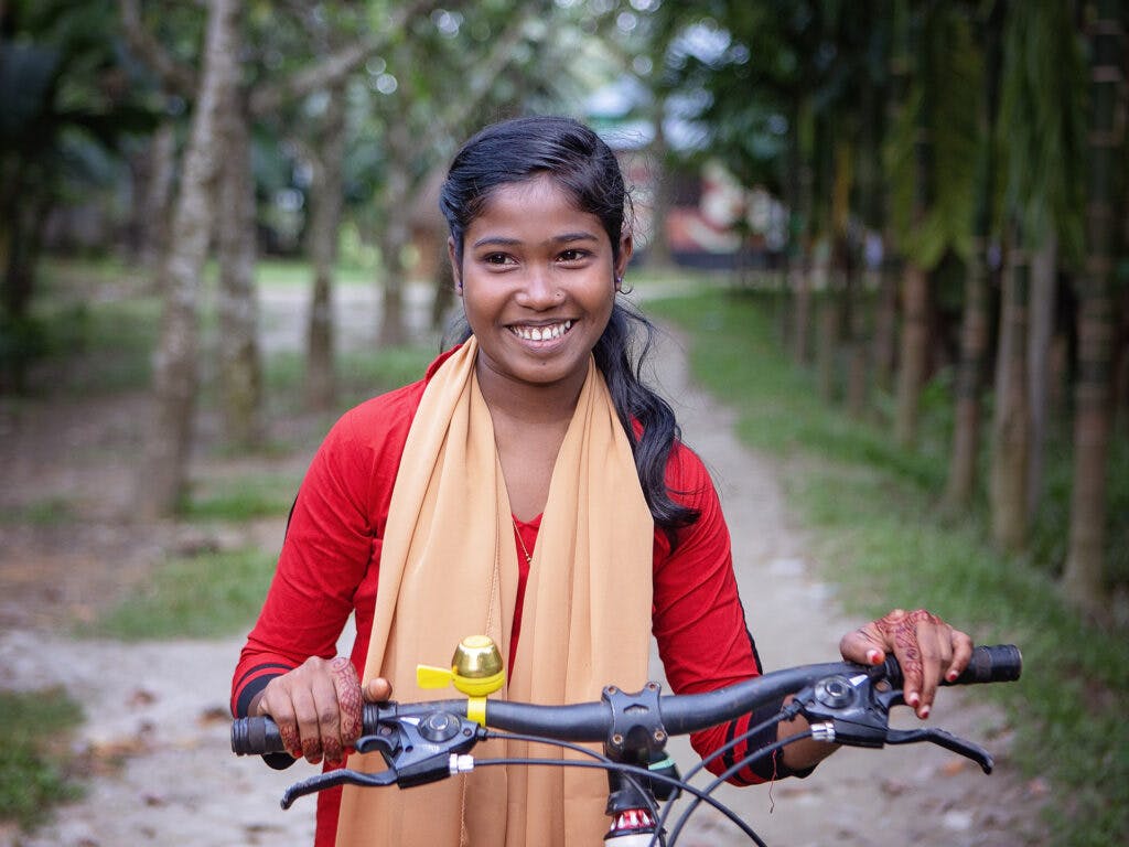 En flicka med röd klänning och gul sjal står framför en cykel och ler.