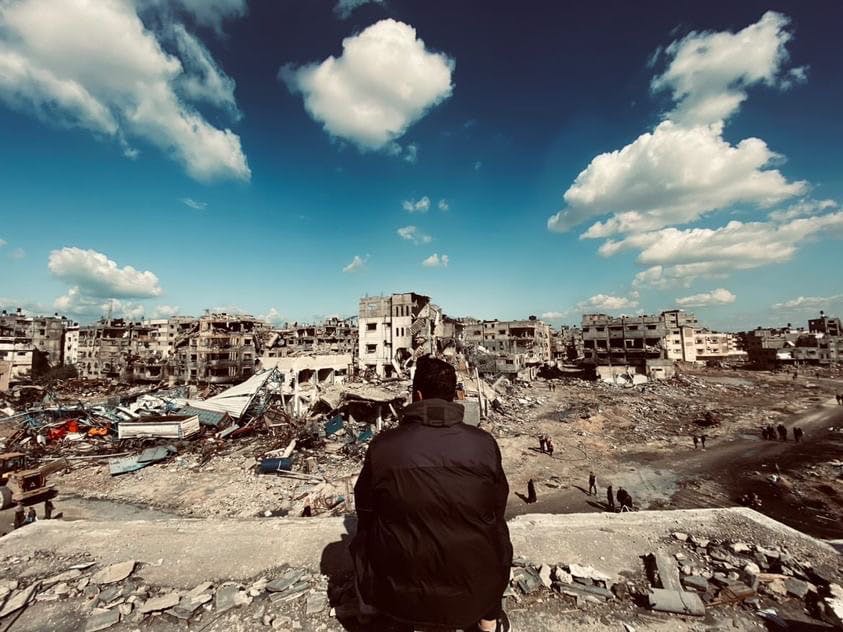 En man sitter i ruiner av bombade byggnader med ryggen mot kameran.