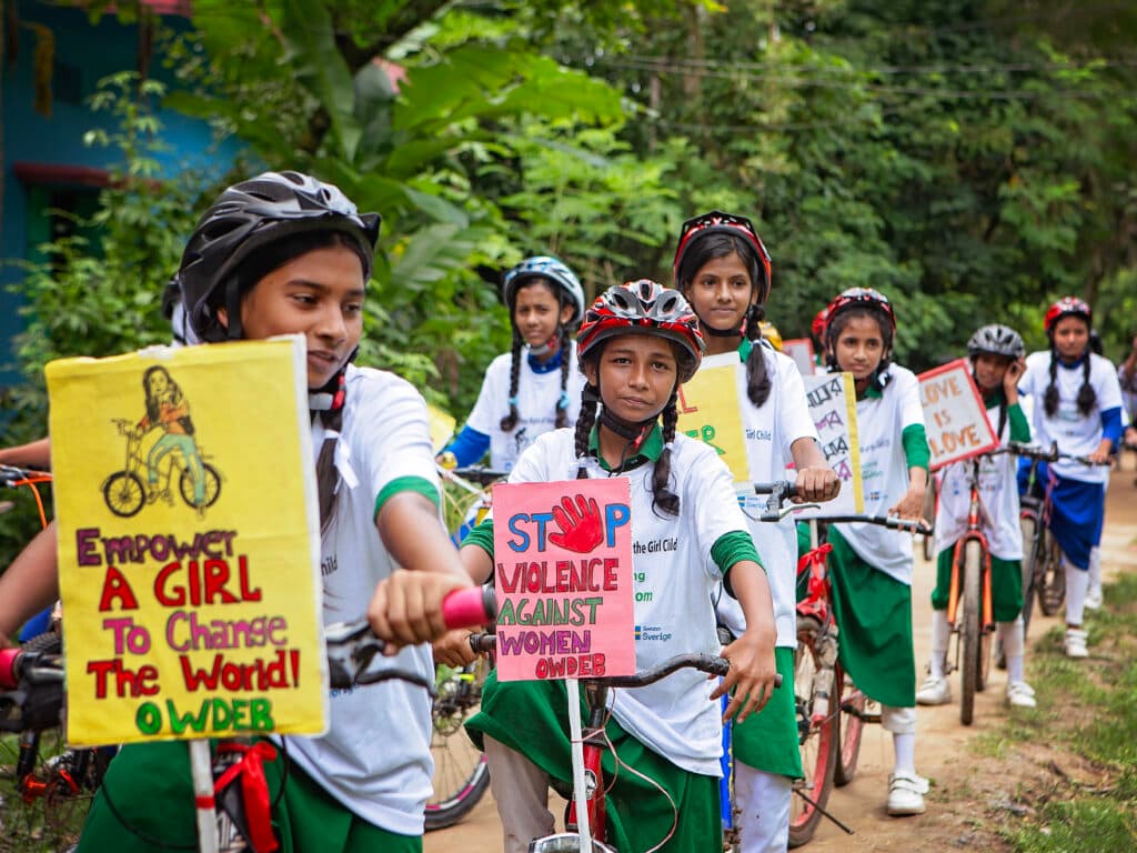 Foto: Diakonia En rad av flickor i skoluniformer. Flickorna står med cyklar som har affischer med budskap för flickors rättigheter.
