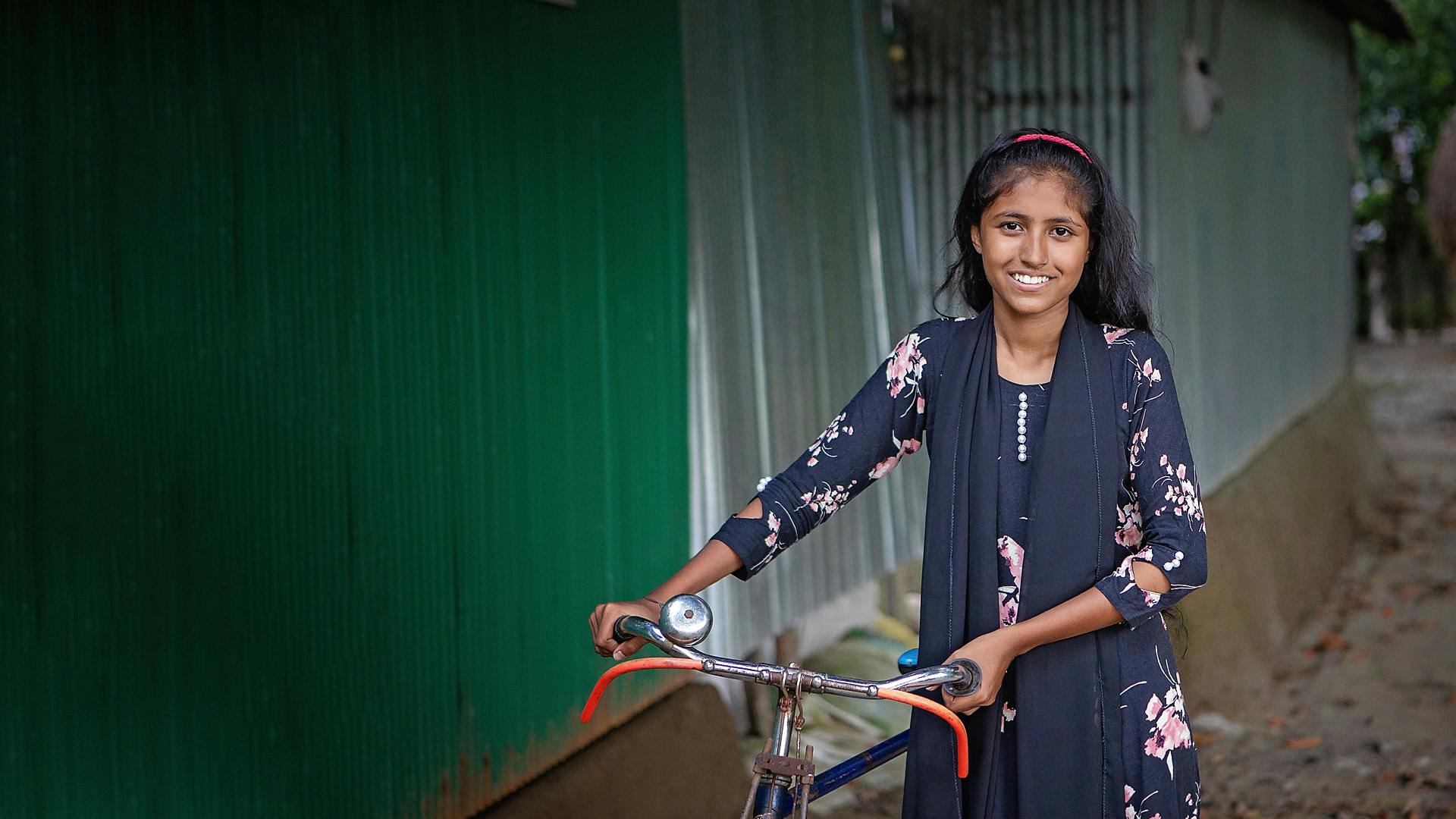En flicka i blommönstrad klänning står med en cykel och ler mot kameran.
