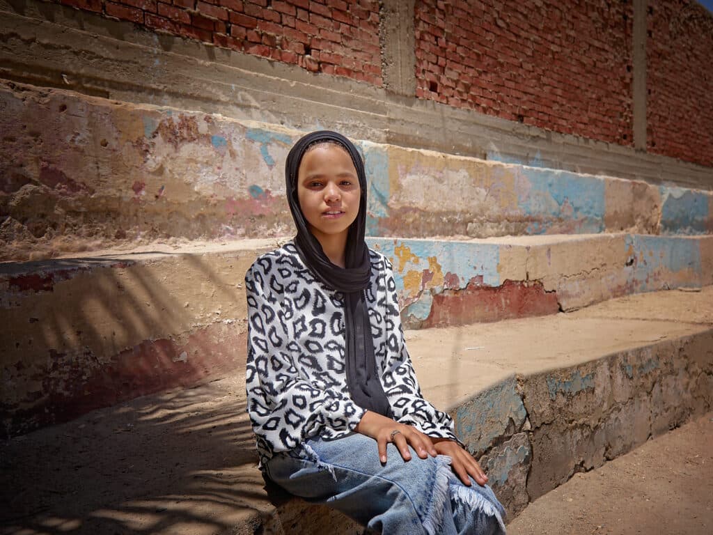 En flicka med svart sjal över huvuet sitter i en stentrappa och ser in i kameran.