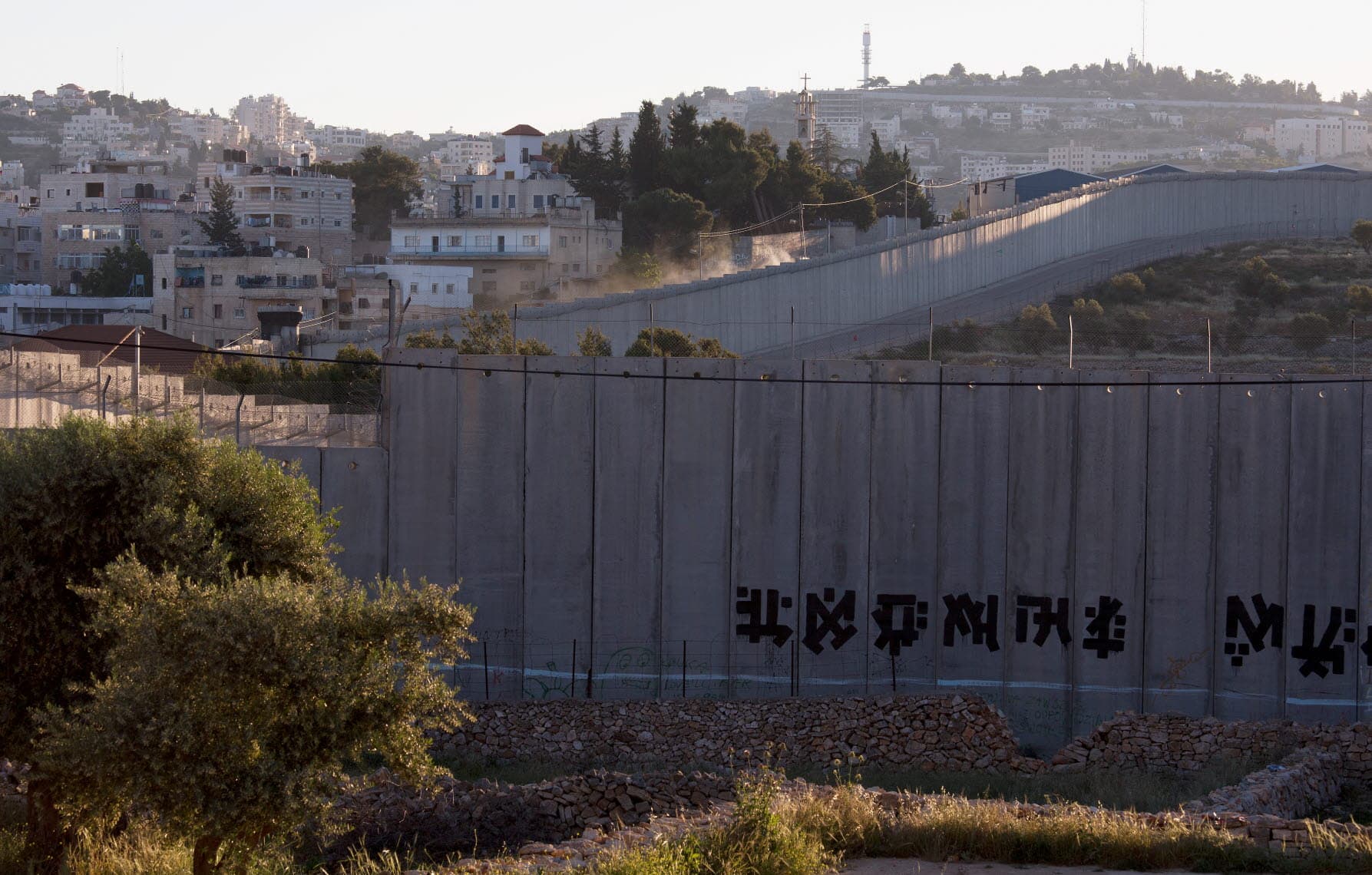 A high concrete wall runs over hills, with houses next to it.