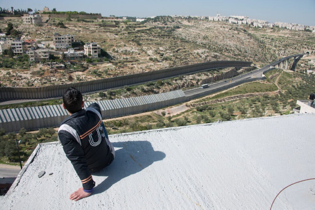 A person on a rooftop overlooking the high concrete wall and settlements on a hill.