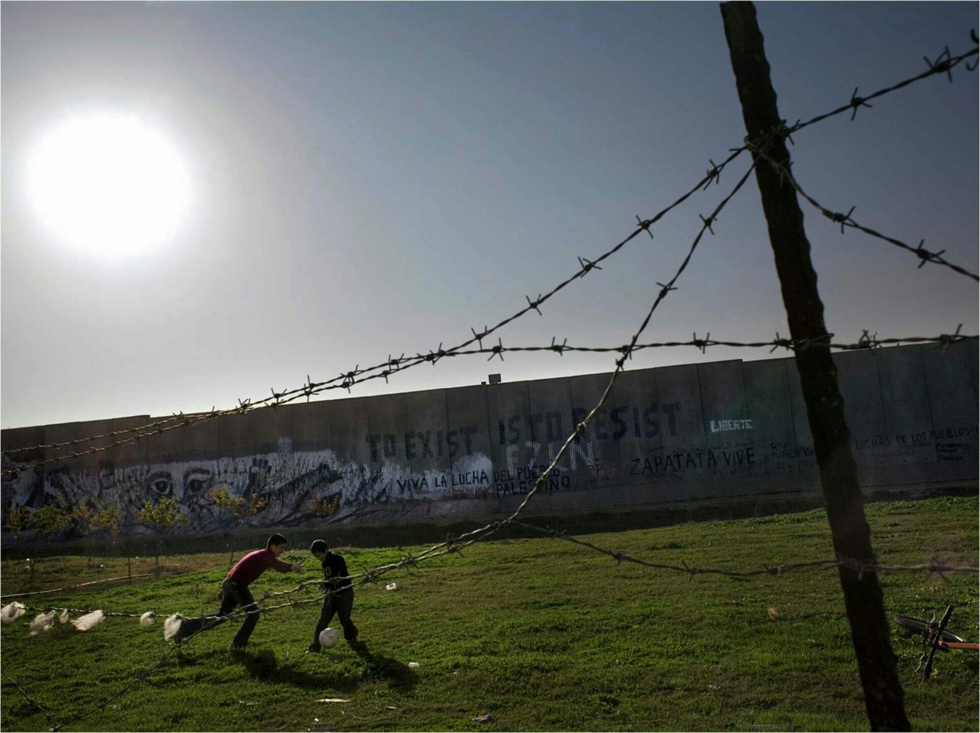 Two boys playing football between barbed wire and a concrete wall.