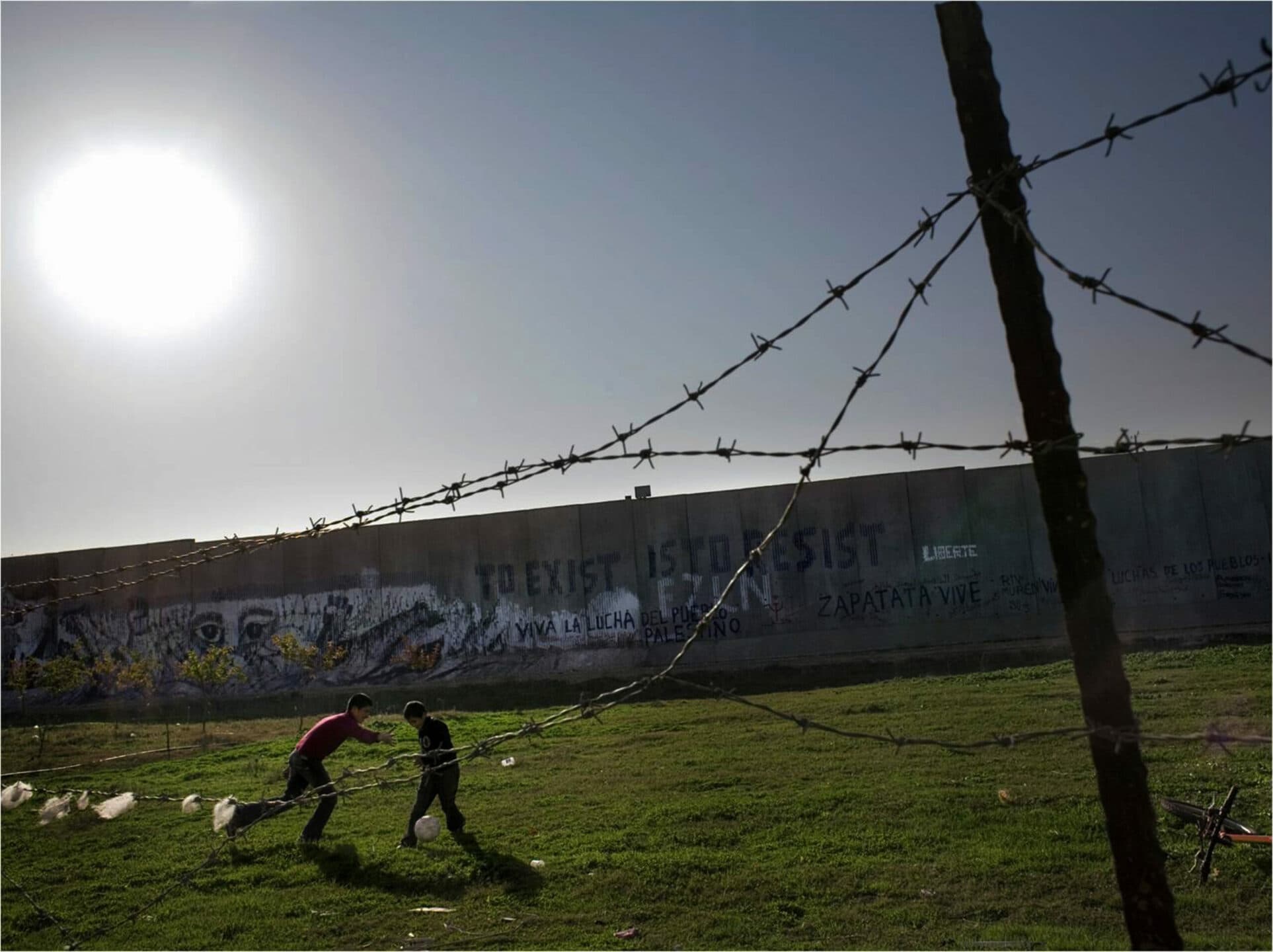 Two boys playing football between barbed wire and a concrete wall.