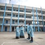 Health workers in protective gear discussing with each other, and in the background a tall building.
