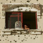 A boy looking out of a window of a building with gun shots on the facade.