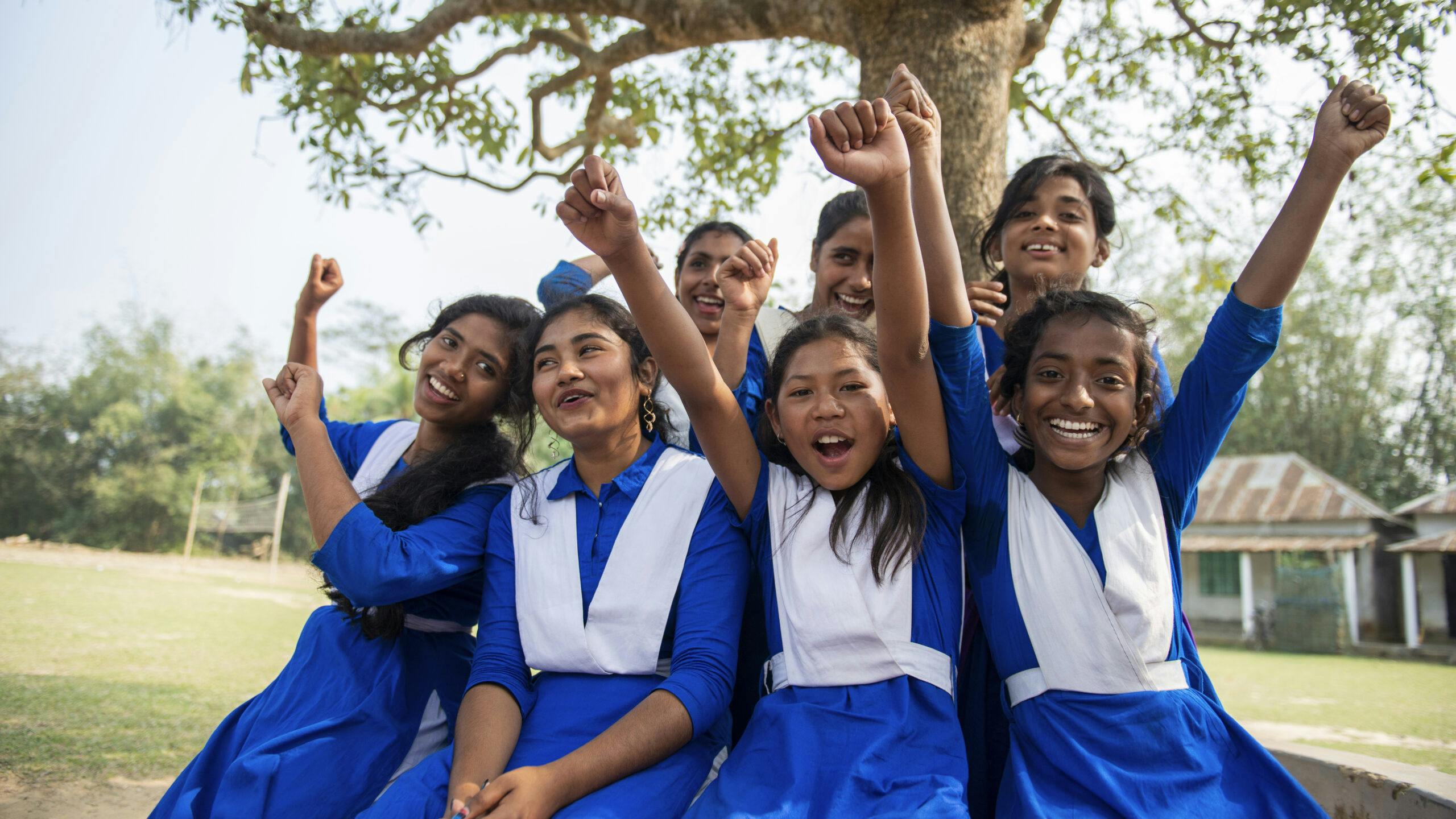 A group of girls in school uniforms sitting next to a tree. They are lifting their hands up.