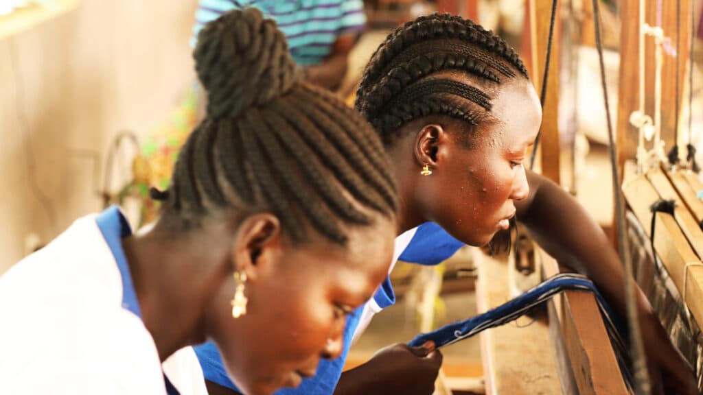 Closeup of two women sitting by a loom, weaving.