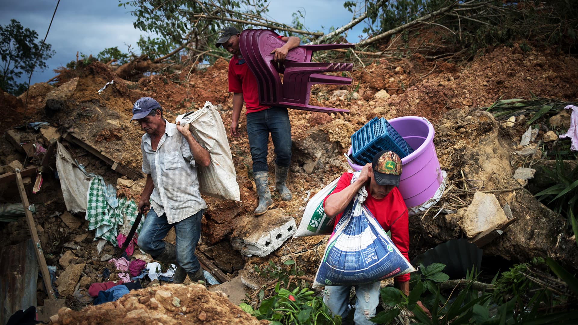 Three men walking down a hill, carrying bags, chairs and other large objects.