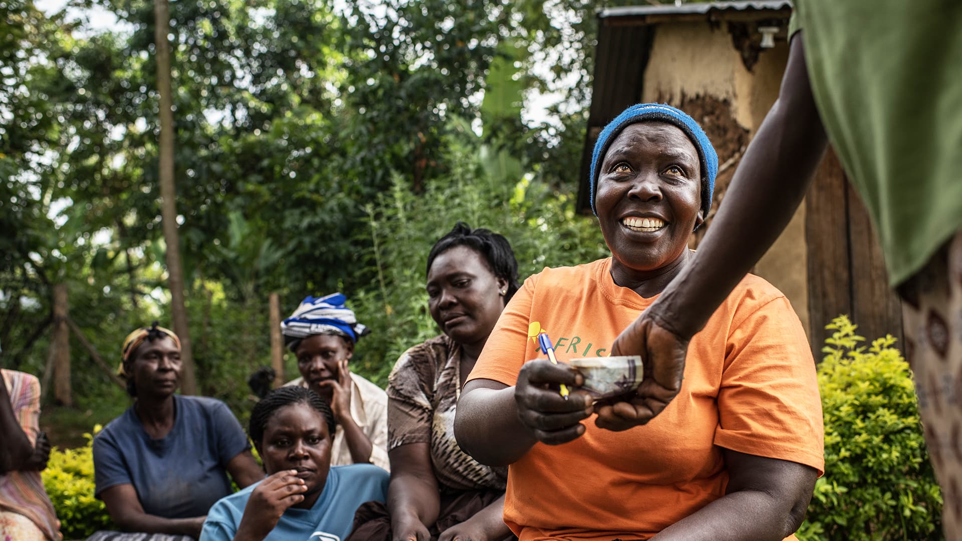 A hand reaching to a woman, giving her money. In the background, there are more women, and trees.