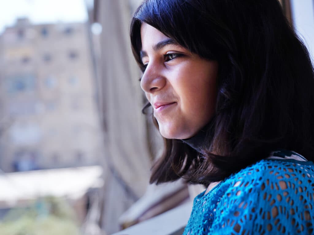 A girl looking out of a window, in the background there are tall grey buildings.