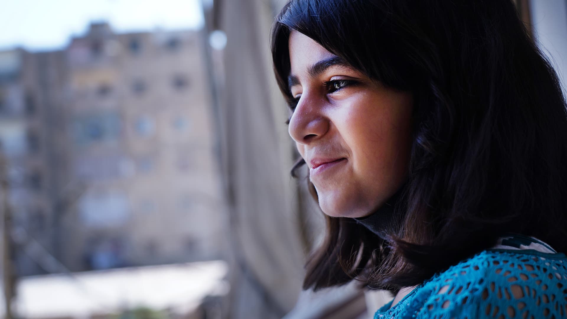 A girl looking out of a window. In the background, there are tall grey buildings.