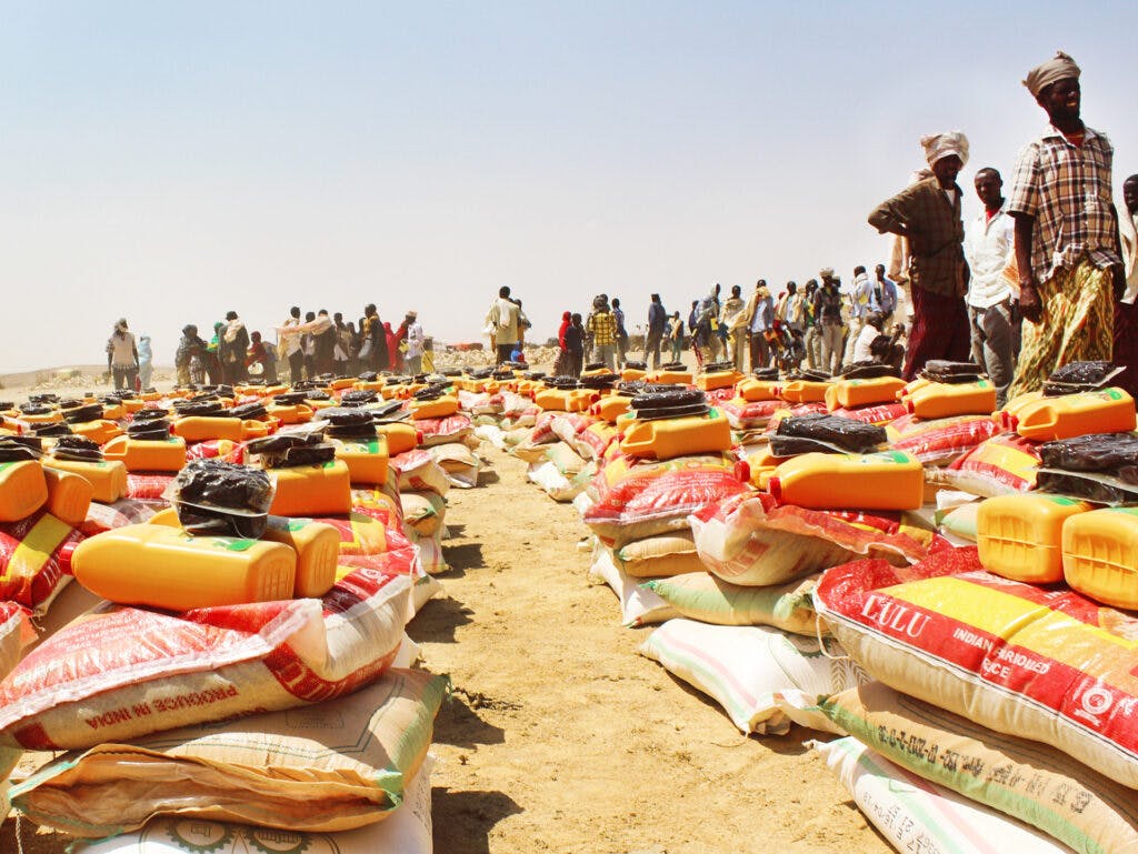 Large bags of food lying in a desert. In the background there is a lot of people.