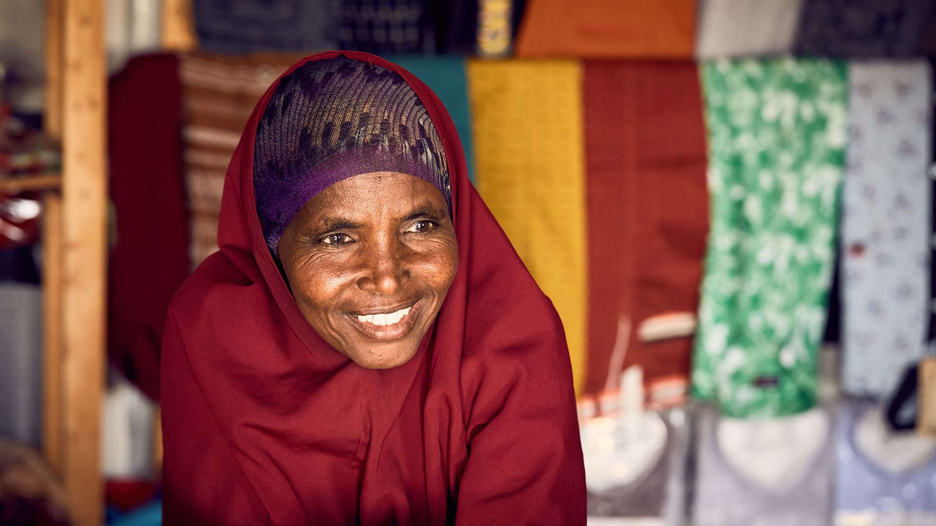 Portrait of a woman in a red hijab. In the background there are fabrics hanging.