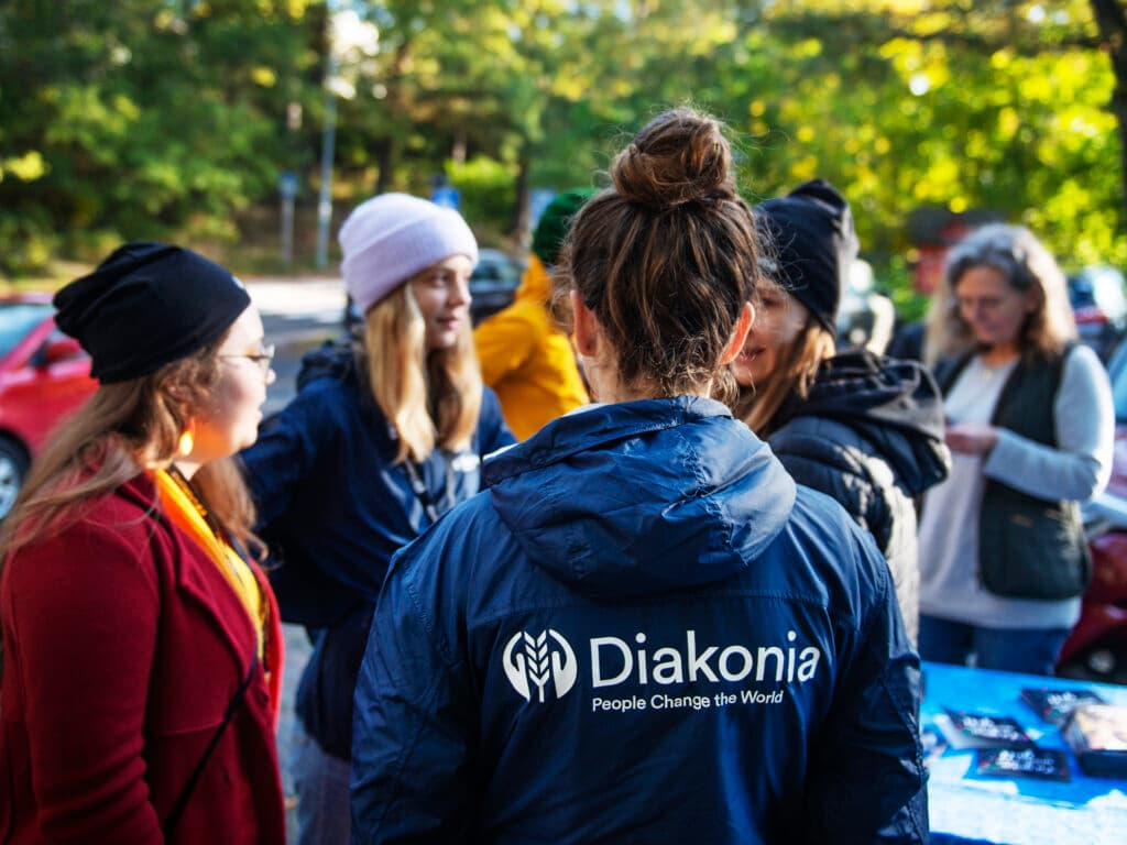 A petrson standing with their back against the camera wearing a Diakonia jacket. In the background there are more people.