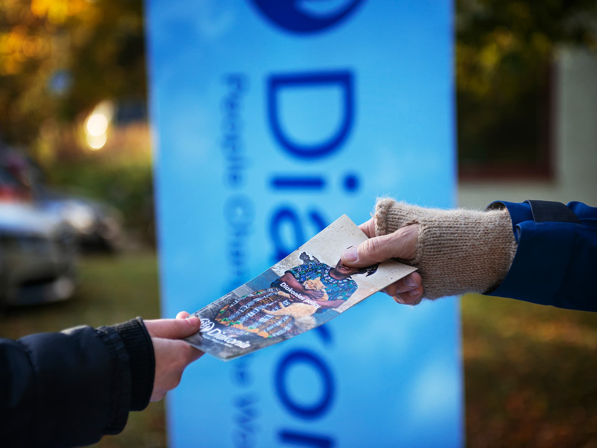 One hand reaching a Diakonia brochure to another hand. In the background there is a large Diakonia sign.