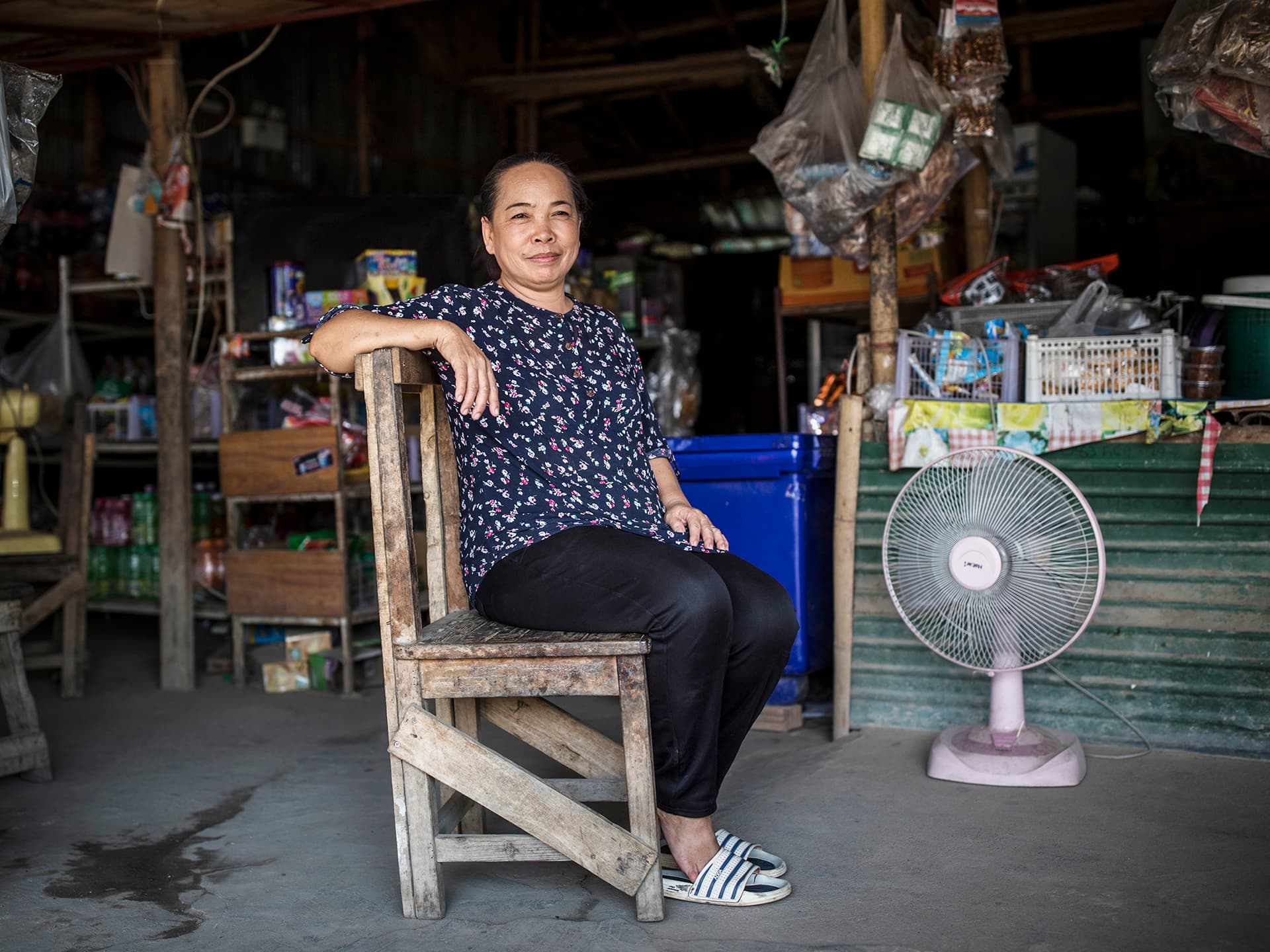 A woman sitting in a chair in an open room. In the background there is a fan and some shelves.