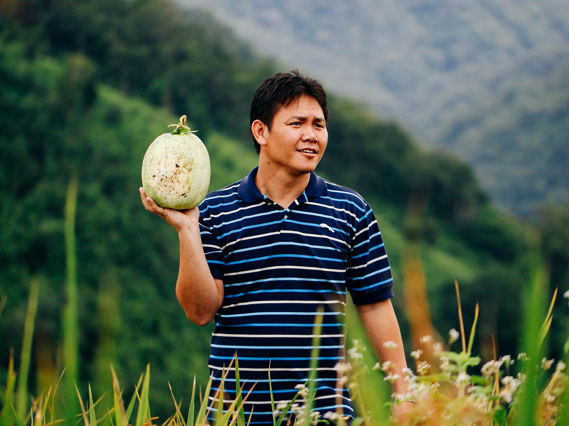 A man standing in a large dield with mountains in the background, holding a large fruit.