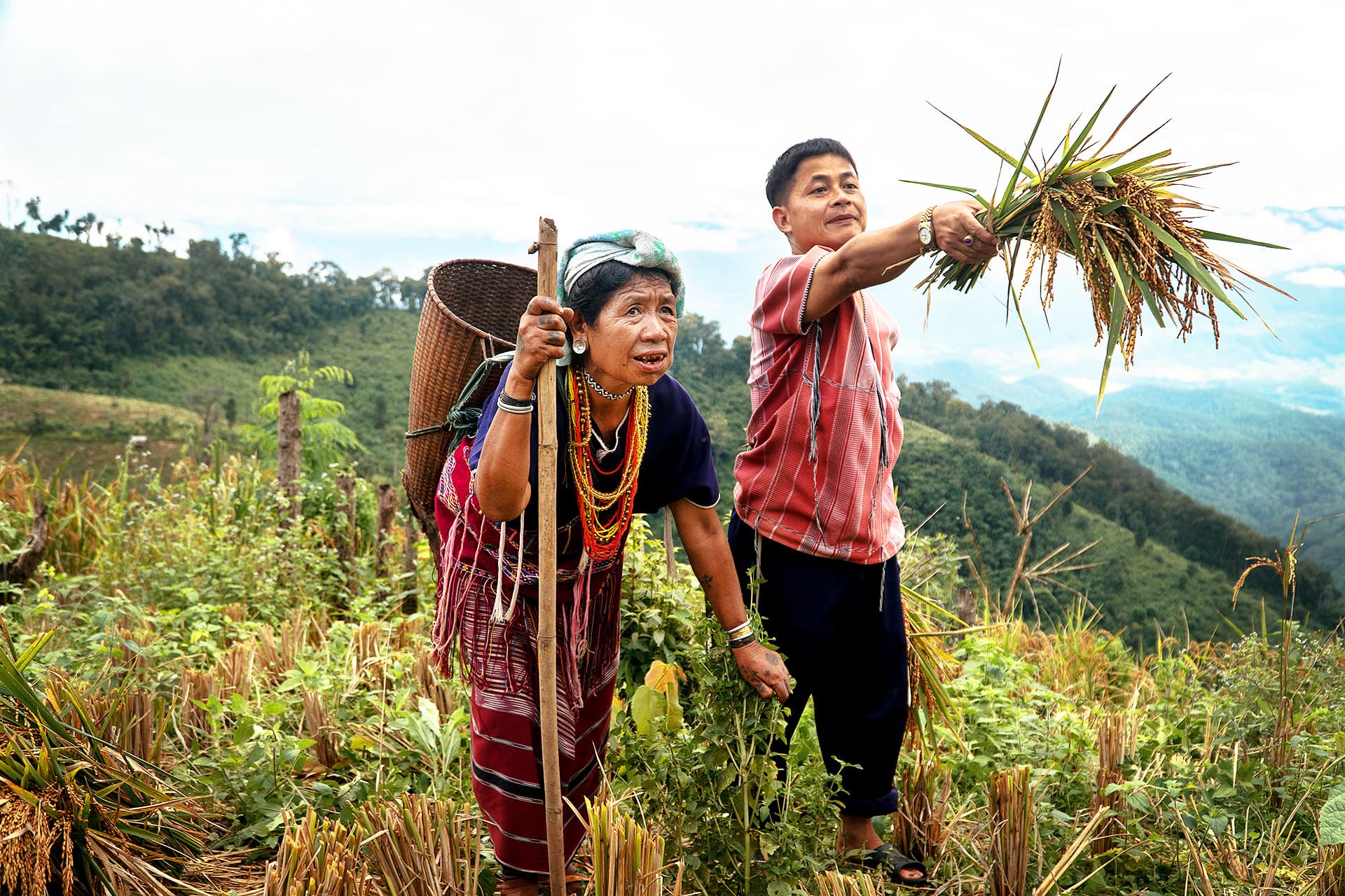 An older woman and a man standing in a field. The woman is wearing a traditional backpack.