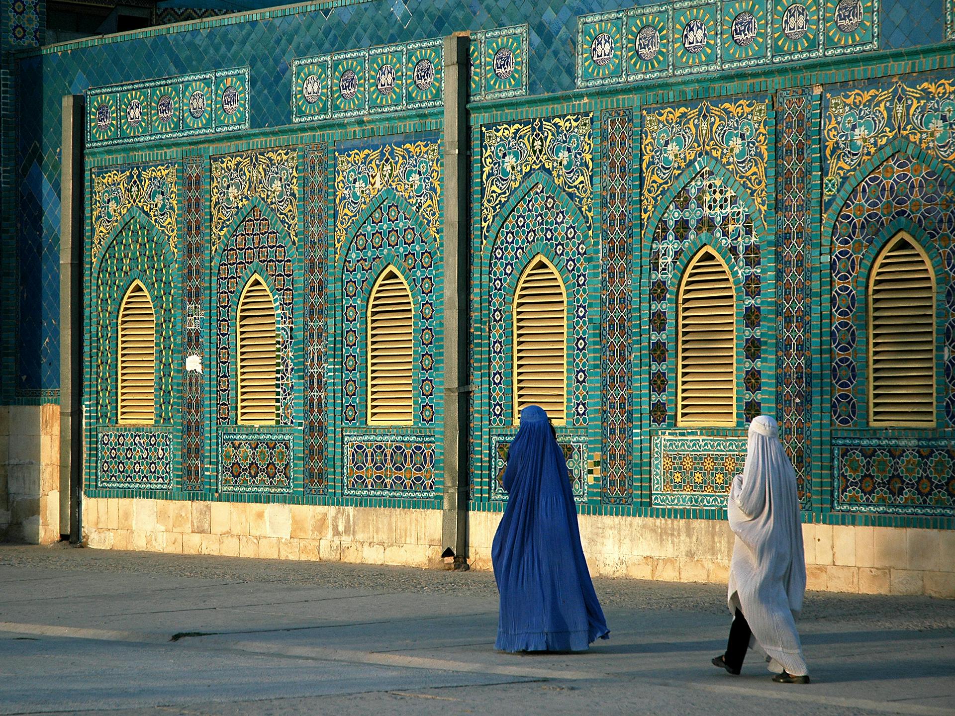 Two women wering burqas seen from behind, walking past a mosque.