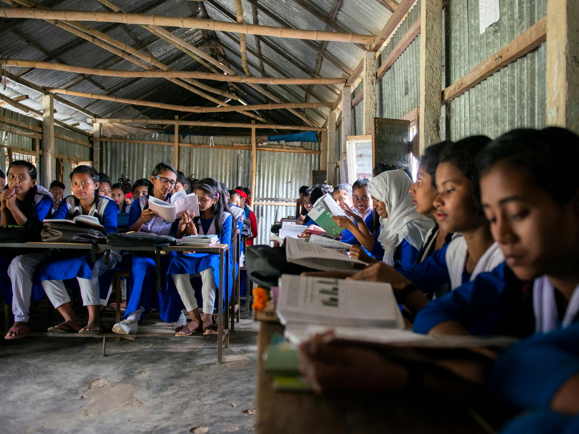 A rural classroom with walls of plate, and a large group of girls sitting by the desks, wearing blue and white school uniforms.