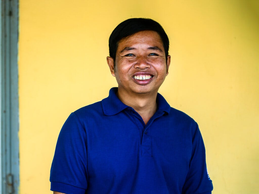 A portrait of a smiling man in a blue t-shirt against a yellow wall.