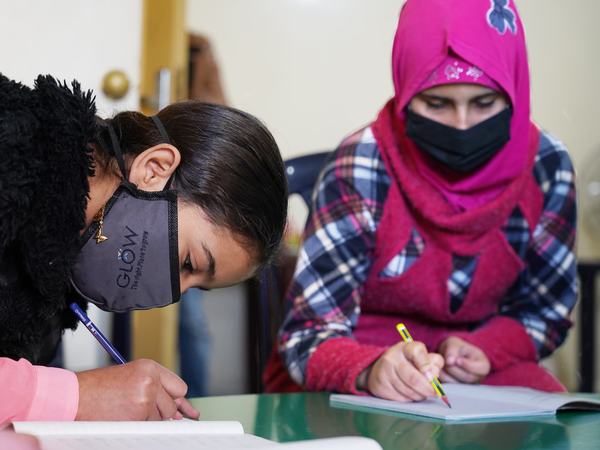 Two girls wearing face masks are leaning down into note books and writing.