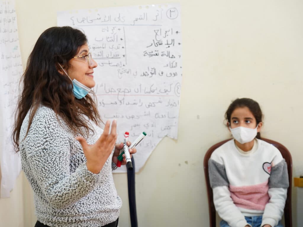 A woman gesticulates with her hand and smiling. Next to her is a girl sitting.