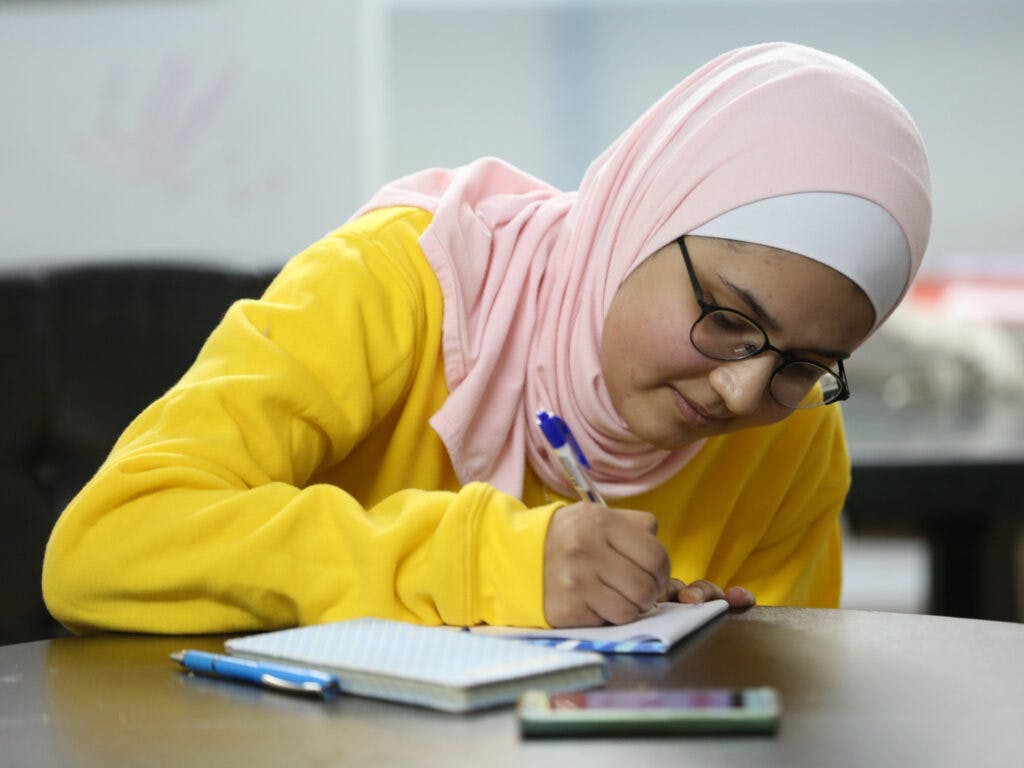 A young woman wearing a pink hijab and a yellow shirt looking down in a book and writing.