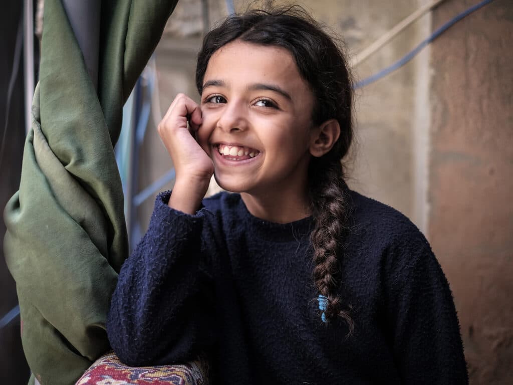 A portrait of a young girl with long dark braided hair, smiling and looking into the camera.