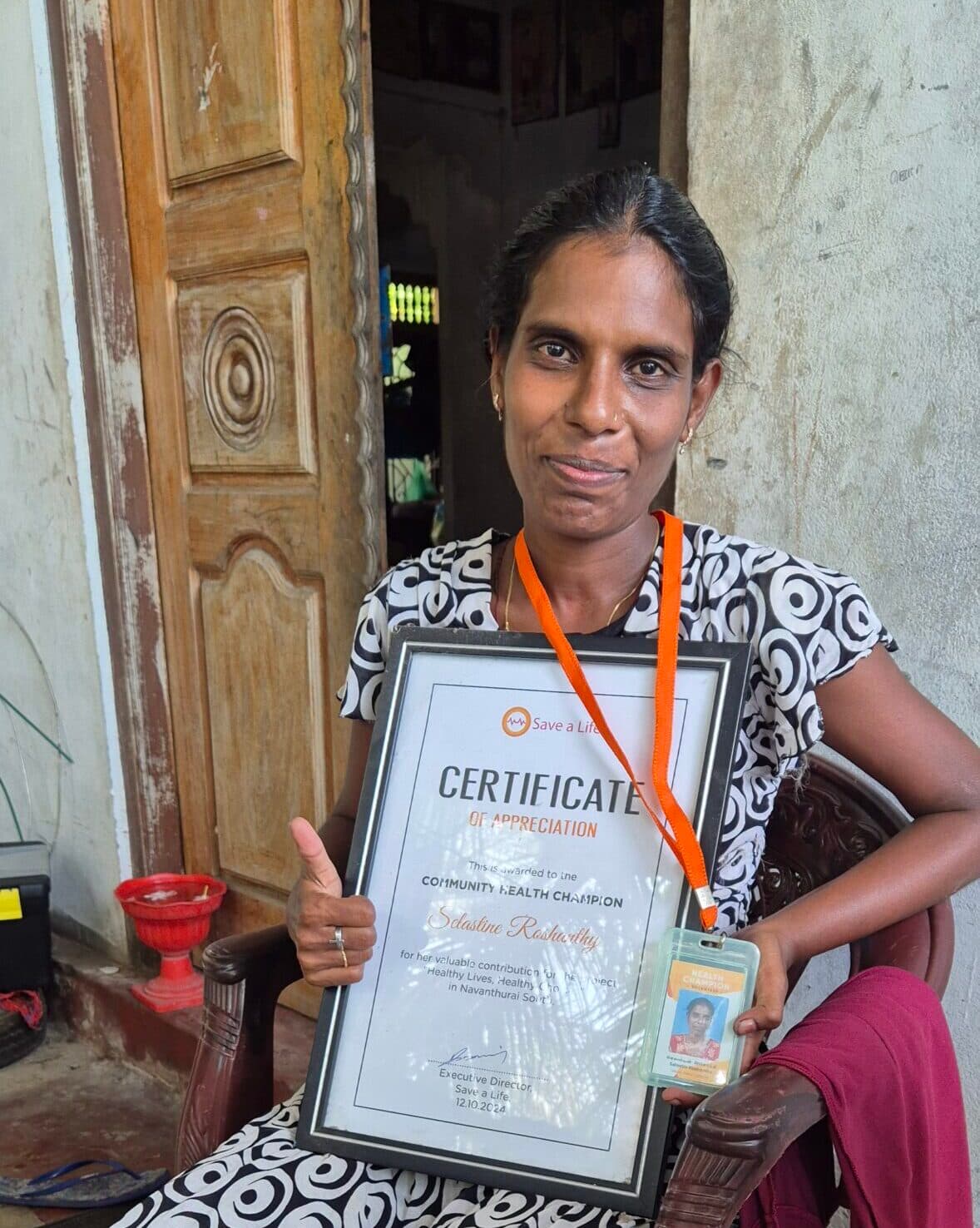 A woman community health champion holding a framed certificate of appreciation, smiling while seated at the entrance of her home.