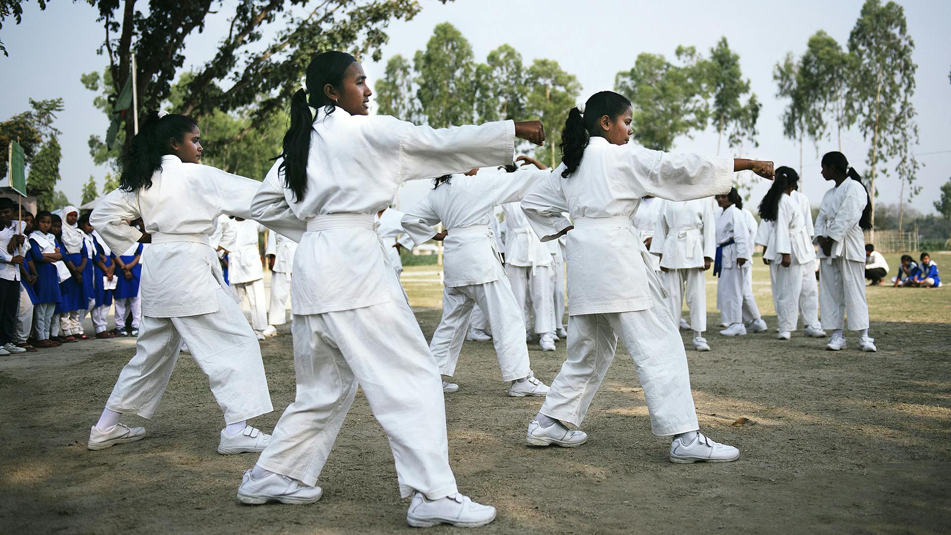 En grupp med unga tjejer som tränar karate utomhus. De har vita karatedräkter på sig. I bakgrunden syns träd och en annan grupp tjejer som tittar på.