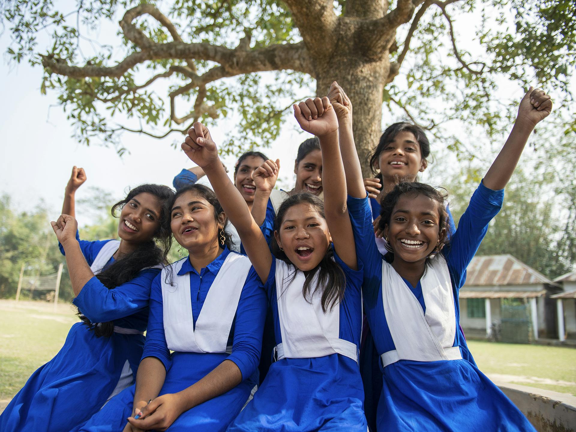 En grupp av leende och skrattande flickor sitter intill ett träd. De är klädda i likadana blåa skoluniformer och sträcker upp händerna i luften.