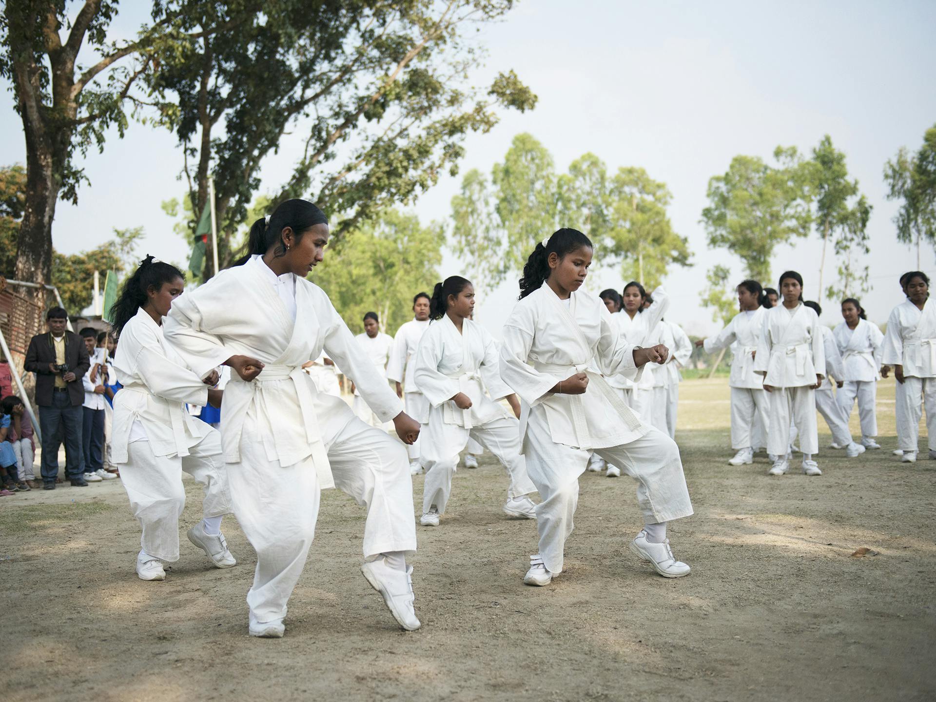 Ett stort antal flickor i vita karatedräkter står utomhus och tränar karate. Bakom dom syns träd och flera personer som tittar på.