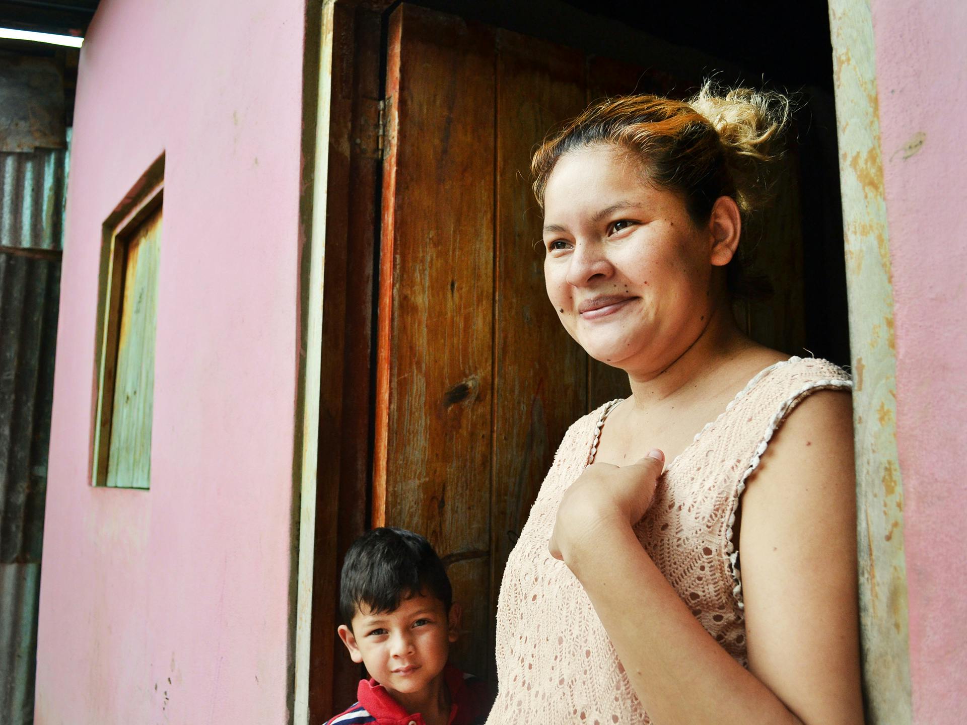 En leende kvinna står i dörröppningen till ett rosa hus. Bredvid henne står ett barn.